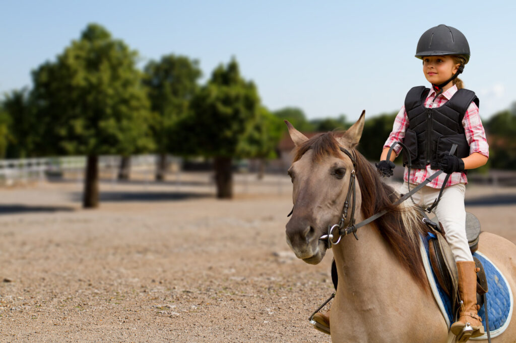 Dziewczynka w kasku i specjalnym stroju do jazdy konnej, jeździ na kucyku na otwartym terenie do jazdy konnej.