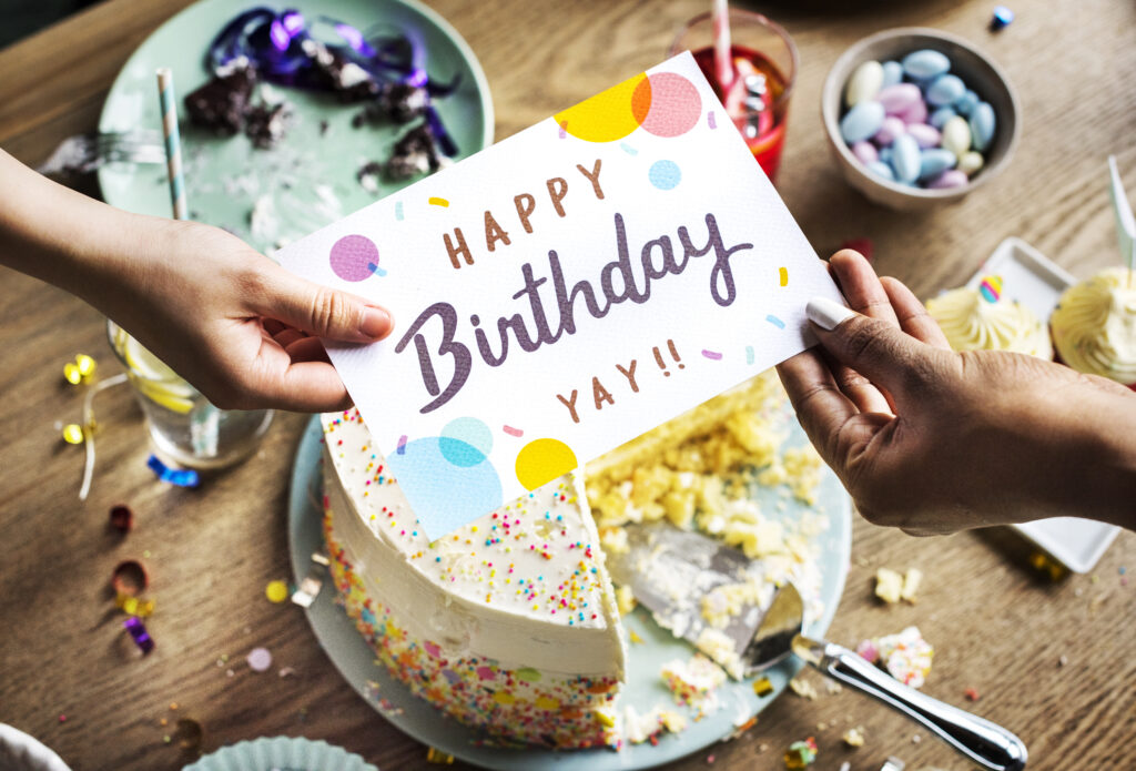 Kartka z napisem ,,happy birthday" przekazywana z rąk do rąk. W tle stół, na którym znajdują się tort, babeczki, szklanki z napojami i cukierki w miseczce.